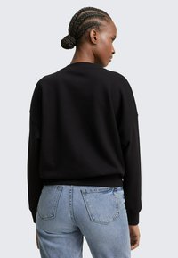 Donna con capelli intrecciati che indossa una maglia nera a maniche lunghe e jeans azzurri chiari, vista di spalle su uno sfondo grigio chiaro uniforme.