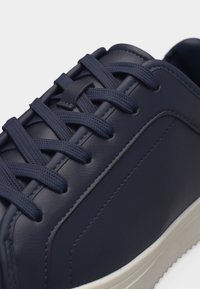 Κοντινό πλάνο ενός αθλητικού παπουτσιού από δέρμα σκούρου μπλε χρώματος με υφασμένες κορδόνια δεμένες μέσω μεταλλικών οπών και λευκή καουτσούκ σόλα ορατή στο κάτω μέρος.