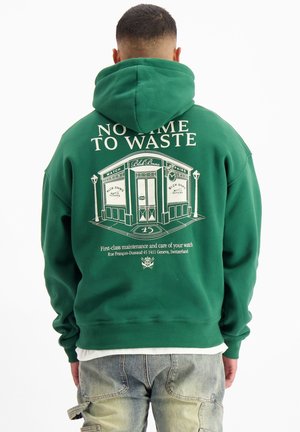 Groene hoodie met een grote witte graphic op de achterkant, featuring een illustratie van een winkel en tekst. Zachte stof, relaxte pasvorm en capuchon ontwerp.