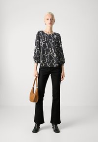 Blusa nera con motivo astratto bianco, maniche lunghe con polsini elasticizzati, abbinata a jeans neri a zampa d'elefante e una borsa color cammello, stivali neri alla caviglia.