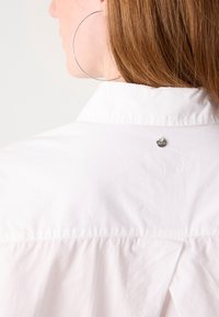 Donna con i capelli rossi che indossa un grande orecchino a cerchio in argento e una camicia bianca con un piccolo logo metallico rotondo sul colletto della parte superiore della schiena.