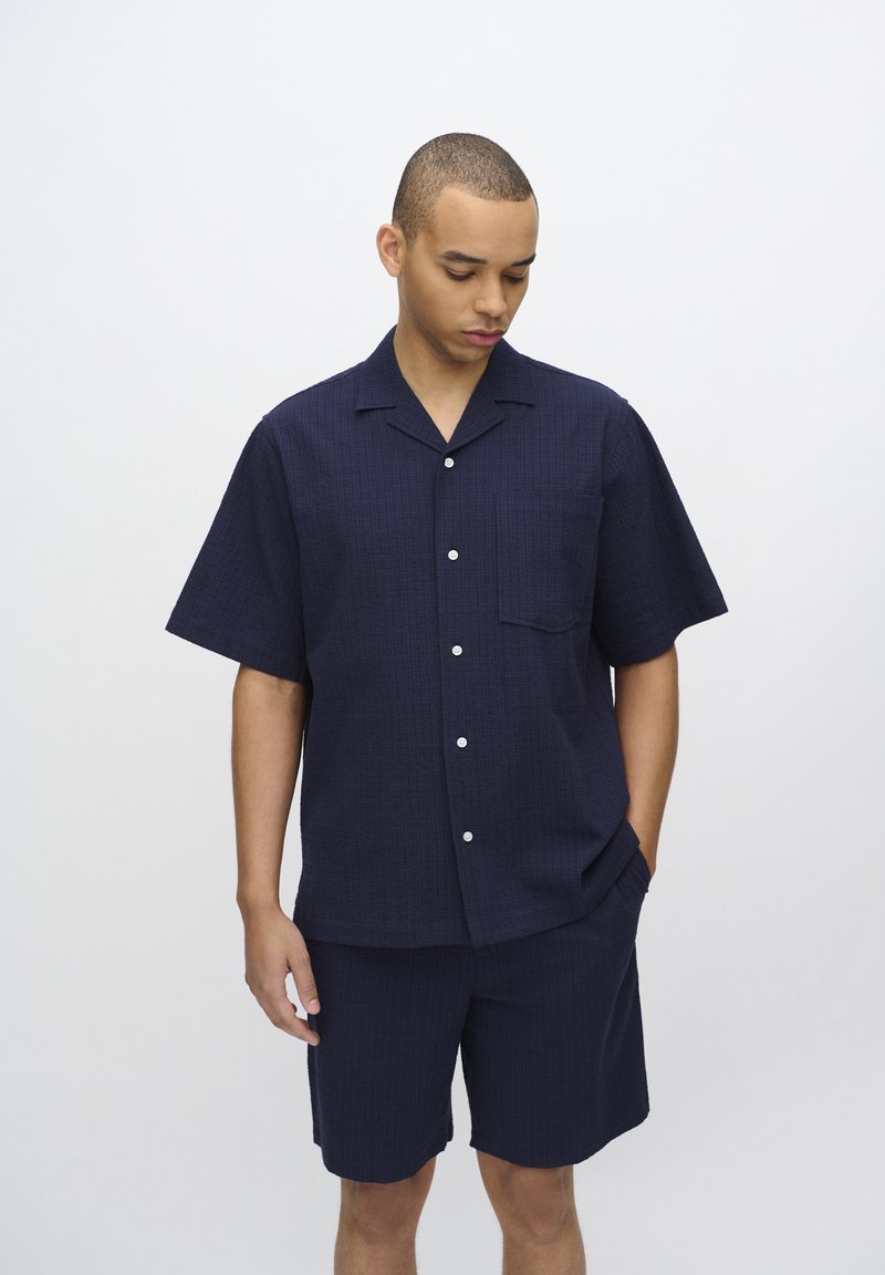 Navy blauw kortemouwenshirt met een gestructureerd patroon en een borstzak, gecombineerd met bijpassende shorts. Bevat witte knopen en heeft een relaxed fit.