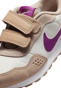 Zapatillas beige con el logo púrpura de Nike, que cuentan con dos correas de velcro, material de ante y malla, detalles perforados y una suela de goma blanca.