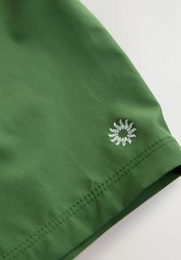 Tissu vert à texture lisse, avec un logo de soleil blanc brodé à l'ourlet. Design simple et finition sur mesure.