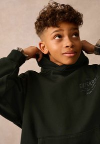 Tmavozelená mikina s kapucňou a prednou vreckom, na hrudi s detailom loga. Má pružné manžety a jemnú textúru.