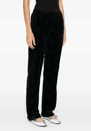 Donna che indossa pantaloni in velluto nero con un sottile motivo a logo su tutta la superficie, abbinati a scarpe piatte argento con punta appuntita.