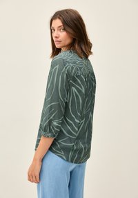 Camicia verde leggera con motivo a foglia, maniche a tre quarti, polsini abbottonati e vestibilità ampia, abbinata a pantaloni azzurri chiari.