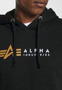 Sweat à capuche noir avec cordons, doté d'un logo en blanc et or très visible : "ALPHA INDUSTRIES" avec un design d'accent angulaire.