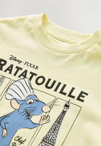 Lysegul t-shirt med grafik af en blå rotte i en kokkehue, med "Ratatouille" tekst og en illustration af Eiffeltårnet.
