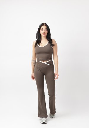 Frau steht, trägt ein eng anliegendes braunes Tanktop und Hosen mit weißer Einfassung, weiße Turnschuhe, neutraler Gesichtsausdruck, schlichter weißer Hintergrund.