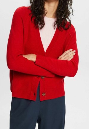 Žena v červeném propínacím svetru přes bílý top se založenýma rukama, ve tmavě modrých kalhotách na jednobarevném pozadí.