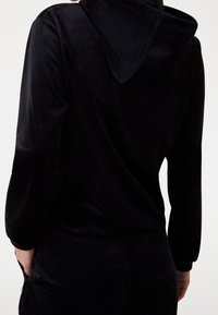 Sweat à capuche en velours noir avec capuche, manches longues et texture lisse. Le design est simple avec une coupe décontractée. Des poches sont visibles.