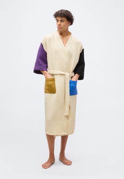 Νεαρός άνδρας φοράει ρόμπα κρεμ με υφή βάφλας, με μωβ και μαύρα μανίκια, μουσταρδί και μπλε τσέπες, στέκεται ξυπόλητος με τα χέρια στις τσέπες.