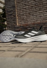 Scarpe da ginnastica nere e grigie con strisce bianche, tomaia in rete e suola testurizzata. Presentano un tallone ammortizzato e un design con lacci.