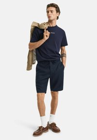 Marine T-shirt met logo, gecombineerd met donkere shorts. Het model draagt bruine schoenen en witte sokken, met een beige jas over één schouder.