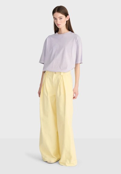 Jeune femme debout portant un t-shirt ample violet clair et un pantalon large jaune pâle sur un fond clair uni.