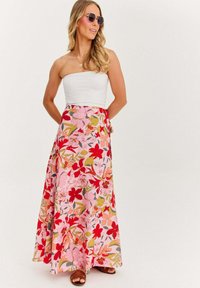 Žena v slnečných okuliaroch, v bielom top s odhalenými ramenami a dlhých ružových sukni s červeným a zeleným kvetinovým vzorom, stojí so založenými rukami za chrbtom.