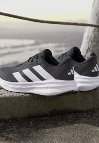 Chaussures de course noires et blanches avec trois bandes blanches sur les côtés, placées sur un rebord en pierre avec un arrière-plan extérieur flou.