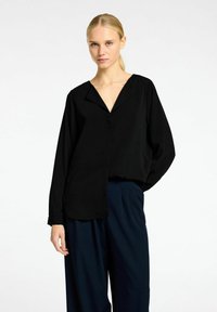 Sort blouse med V-hals, lange ærmer, løs pasform og en glat tekstur, parret med marineblå bukser. Enkel design med minimal detaljer.