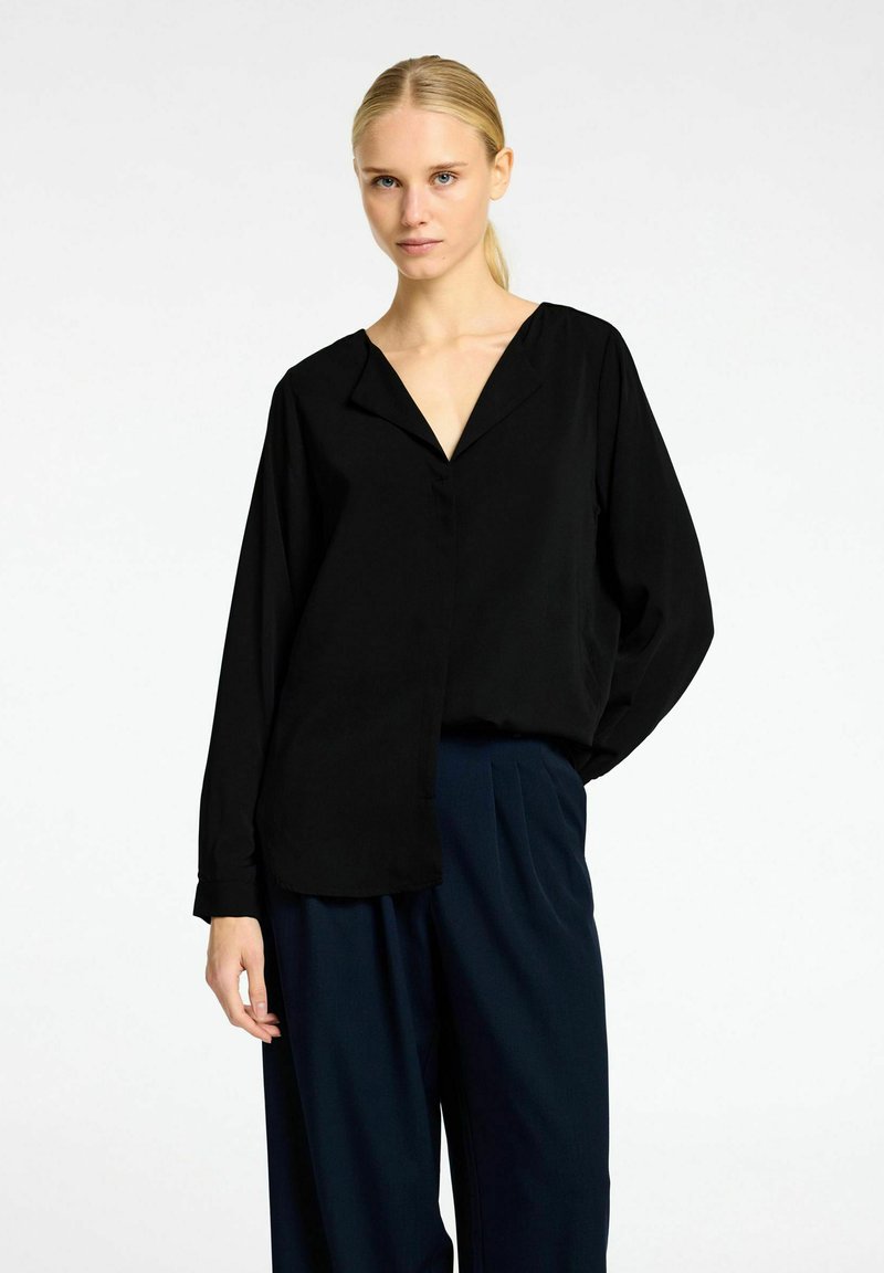 Sort blouse med V-hals, lange ærmer, løs pasform og en glat tekstur, parret med marineblå bukser. Enkel design med minimal detaljer.