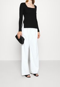 Femme portant un haut noir à manches longues et décolleté carré, un pantalon blanc à jambes larges, des sandales à talons noires et tenant un sac à main noir.