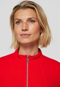 Red zip-up jacket with a textured pattern, high collar, and metallic zipper. The fabric appears soft and lightweight.