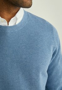 Blauer gestrickter Pullover mit Rundhalsausschnitt, der ein strukturiertes Muster und gerippte Bündchen aufweist. Über einem weißen Hemd mit Kragen getragen.