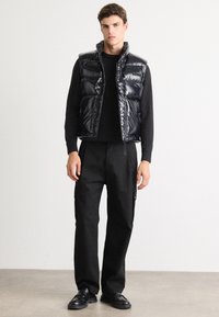 Schwarze, gepolsterte Weste mit glänzender Oberfläche, Reißverschluss vorne und hohem Kragen. Getragen über einem schwarzen Langarmshirt und kombiniert mit lockeren schwarzen Hosen.