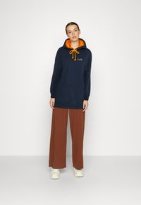 Sudadera azul marino con forro naranja, cordón y bolsillo frontal. Combinada con pantalones de pierna ancha marrones y zapatillas blancas. Diseño sencillo.