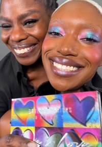 Paletten har fargerike hjerte mønstre med nyanser av rosa, blå og grønn. To personer smiler, og viser frem livlig sminke og glansfulle lepper.