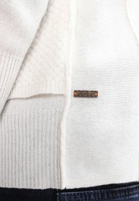 Vit stickad tröja med ribbade och texturerade mönster. Inkluderar en liten rektangulär etikett med texten "DOSS ORANGE" och orange detaljer.