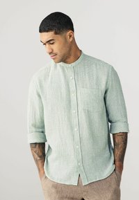 Camisa de color verde claro con cuello mao, tejido texturizado y un solo bolsillo en el pecho. Mangas largas remangadas en los puños.