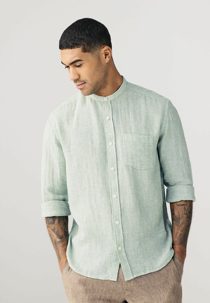 Camisa de color verde claro con cuello mao, tejido texturizado y un solo bolsillo en el pecho. Mangas largas remangadas en los puños.