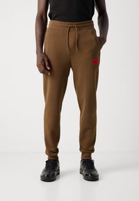 Man wearing brown jogger pants with drawstring and red HUGO logo patch, paired with black sneakers and a black sleeveless top.