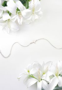 Collana in argento con un piccolo pendente a croce, posata tra fiori bianchi con petali lisci e accenti verdi su uno sfondo chiaro.