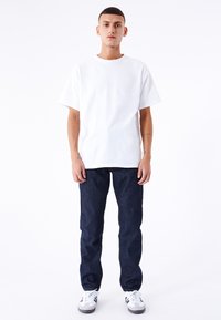 Camiseta blanca de algodón con cuello redondo, combinada con jeans de mezclilla azul oscuro. El modelo lleva puestos unos zapatos deportivos blancos y grises con detalles en negro.