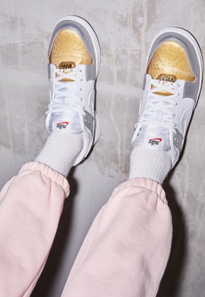 Stopy w białych sneakersach Nike ze złotymi czubkami, białych skarpetkach i jasnoróżowych spodniach na tle szarej betonowej podłogi.