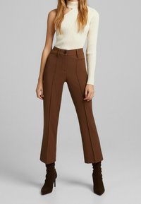 Brązowe, dopasowane spodnie z wysokim stanem i prostymi nogawkami, zestawione z prążkowanym, kremowym topem na jedno ramię oraz pasującymi, wysokimi kozakami na obcasie.