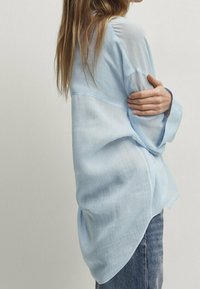 Ljusblå genomskinlig blus med långa ärmar och rundad nederkant. Har en lös passform och mjuk textur, bärs över mörka denimjeans.