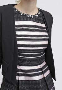 Svart kavaj över en randig klänning med svarta och rosa horisontella ränder, samt en smyckad halsringning med texturerade tygdetaljer.