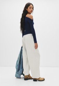 Mörkblå off-shoulder topp, ljusgrå vida byxor, beiga ankelstövlar med spännen, håller i en jeansjacka. Släta tygtexturer.