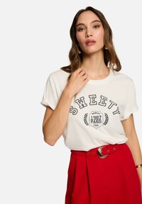 T-shirt en coton blanc avec le texte "SWEETY" et un design graphique ; manches courtes roulées ; associé à un pantalon rouge taille haute et une ceinture métallique.