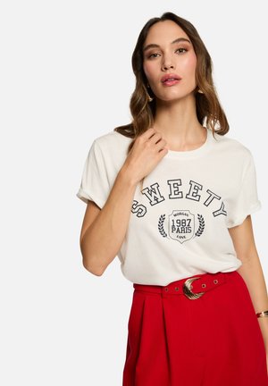 T-shirt en coton blanc avec le texte "SWEETY" et un design graphique ; manches courtes roulées ; associé à un pantalon rouge taille haute et une ceinture métallique.