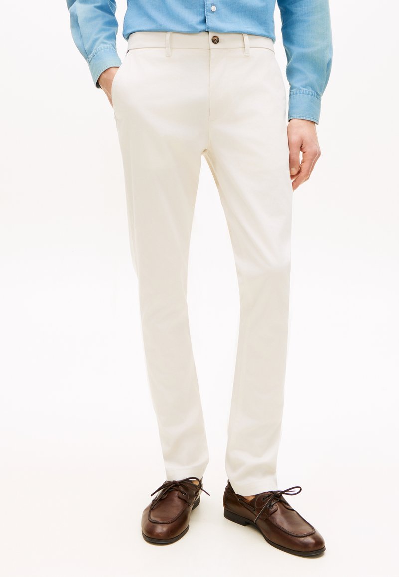 Άνδρας που φοράει λευκό στενό παντελόνι, γαλάζιο πουκάμισο μαζεμένο μέσα, και καφέ δερμάτινα παπούτσια με κορδόνια, στέκεται με τα χέρια στις τσέπες.