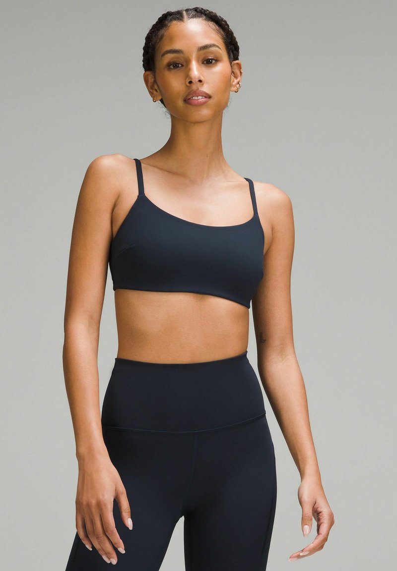 Čierny športový podprsenka s tenkými ramienkami, vyrobená z hladkého materiálu. Má zaoblený výstrih a skrátený strih, doplnená vysokými čiernymi legínami.