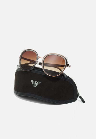 Per didelės apimties saulės akiniai su gradientiniais rudiuose lęšiais ir skaidriais rėmeliais, resting on a black soft case with a silver logo.