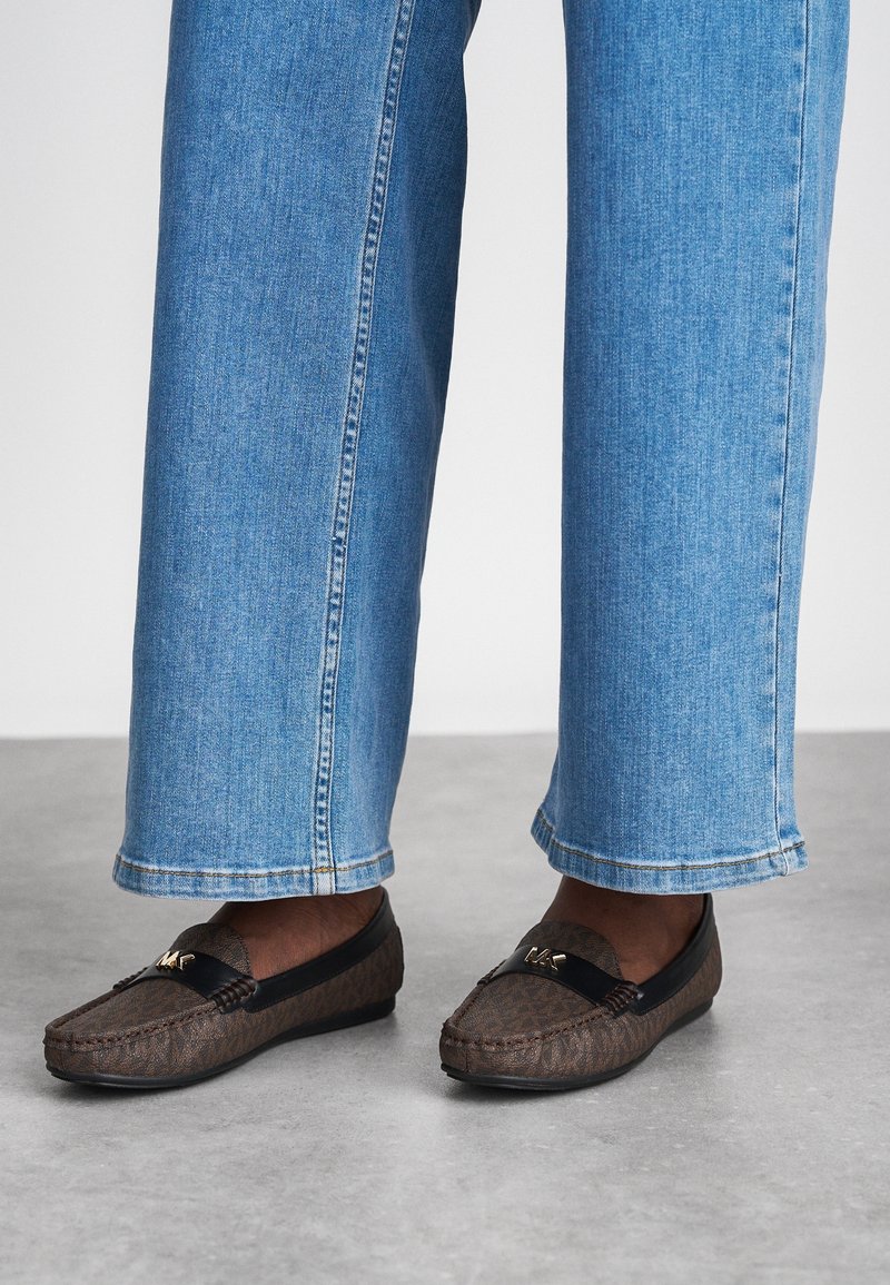 Personne portant des mocassins marron à motifs, avec une bordure noire et un détail doré, associés à un jean bleu clair à coupe droite.