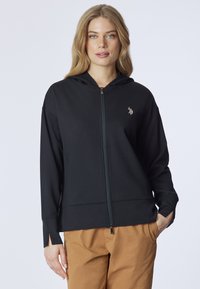 Felpa nera con zip realizzata in tessuto morbido, dotata di cappuccio, spacchi laterali e un piccolo logo decorativo. Abbinata a pantaloni color sabbia.