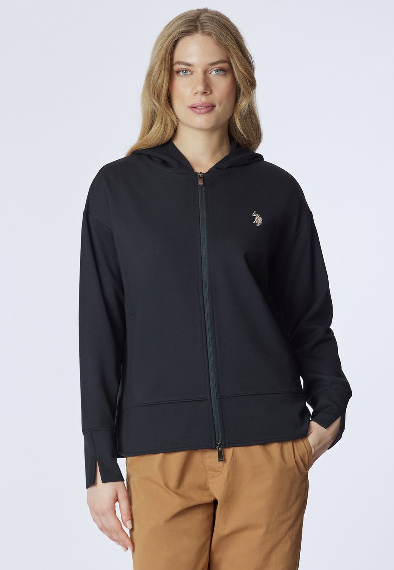 Felpa nera con zip realizzata in tessuto morbido, dotata di cappuccio, spacchi laterali e un piccolo logo decorativo. Abbinata a pantaloni color sabbia.