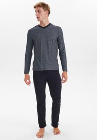 Grå tekstureret bluse med lange ærmer og sort V-hals samt sorte tætsiddende bukser. Barfodet fremtoning med en minimalistisk stil.